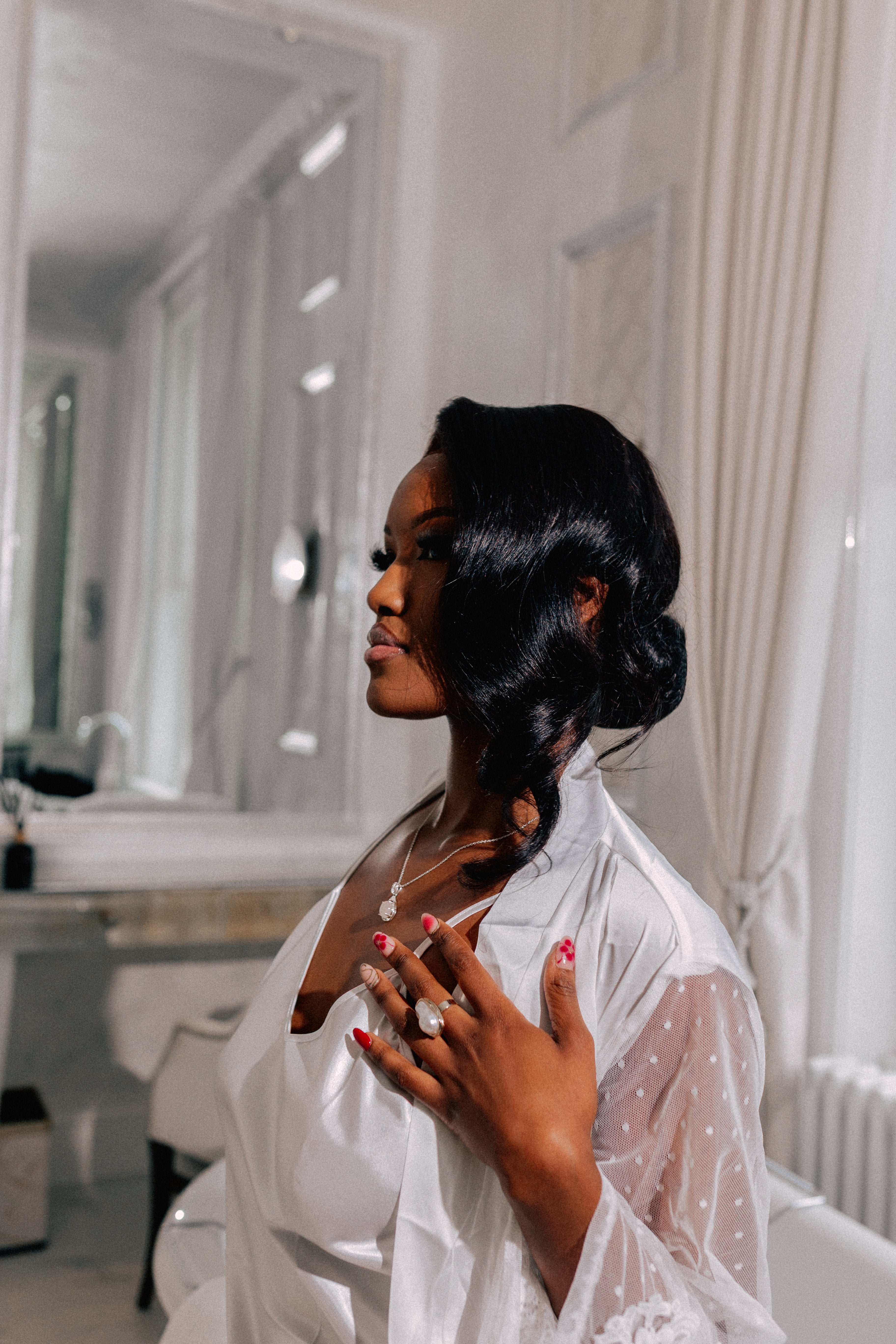 *"Elegant Black woman with vintage Hollywood waves hairstyle wearing a white satin bridal robe and sheer lace sleeves, showcasing a pearl engagement ring and matching necklace in a luxurious white bathroom setting."